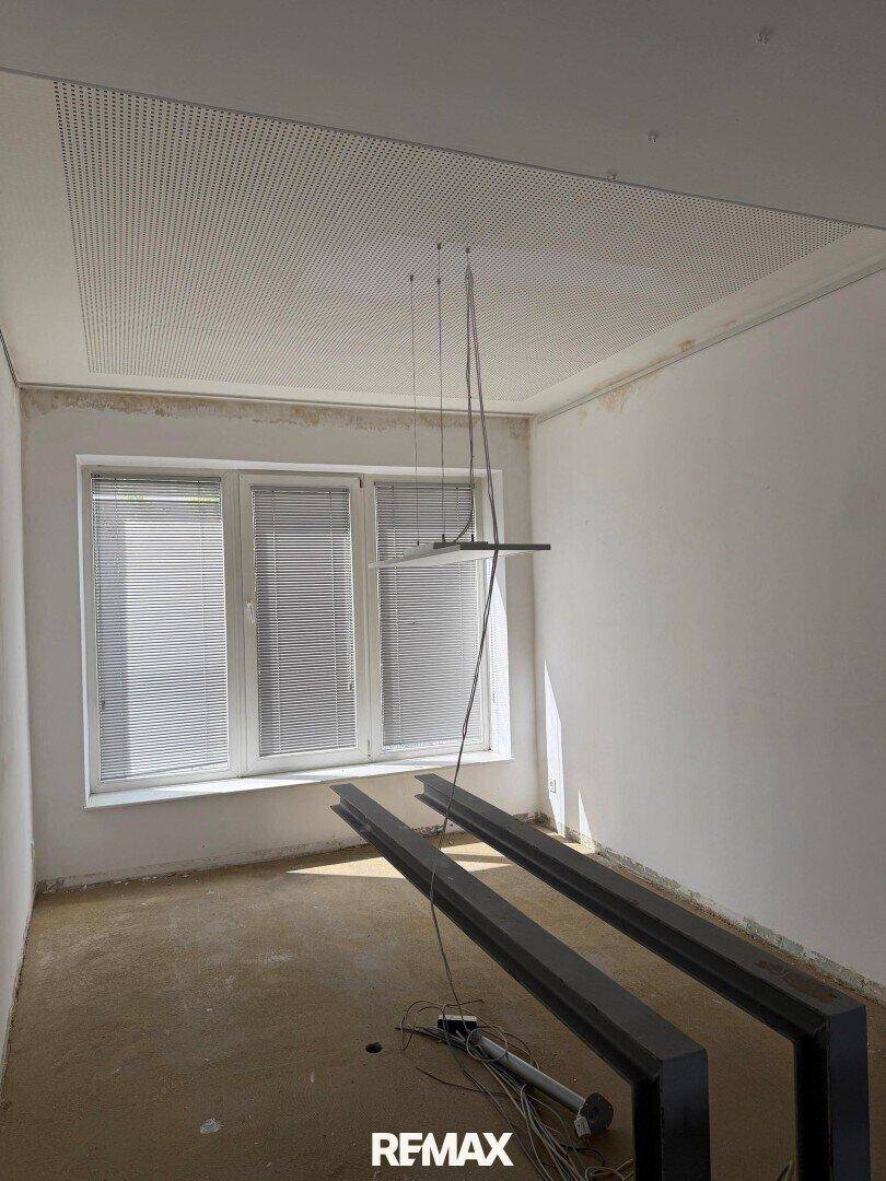Leerer Raum mit großem Fenster, perforierter Decke und sichtbaren Installationskabeln.