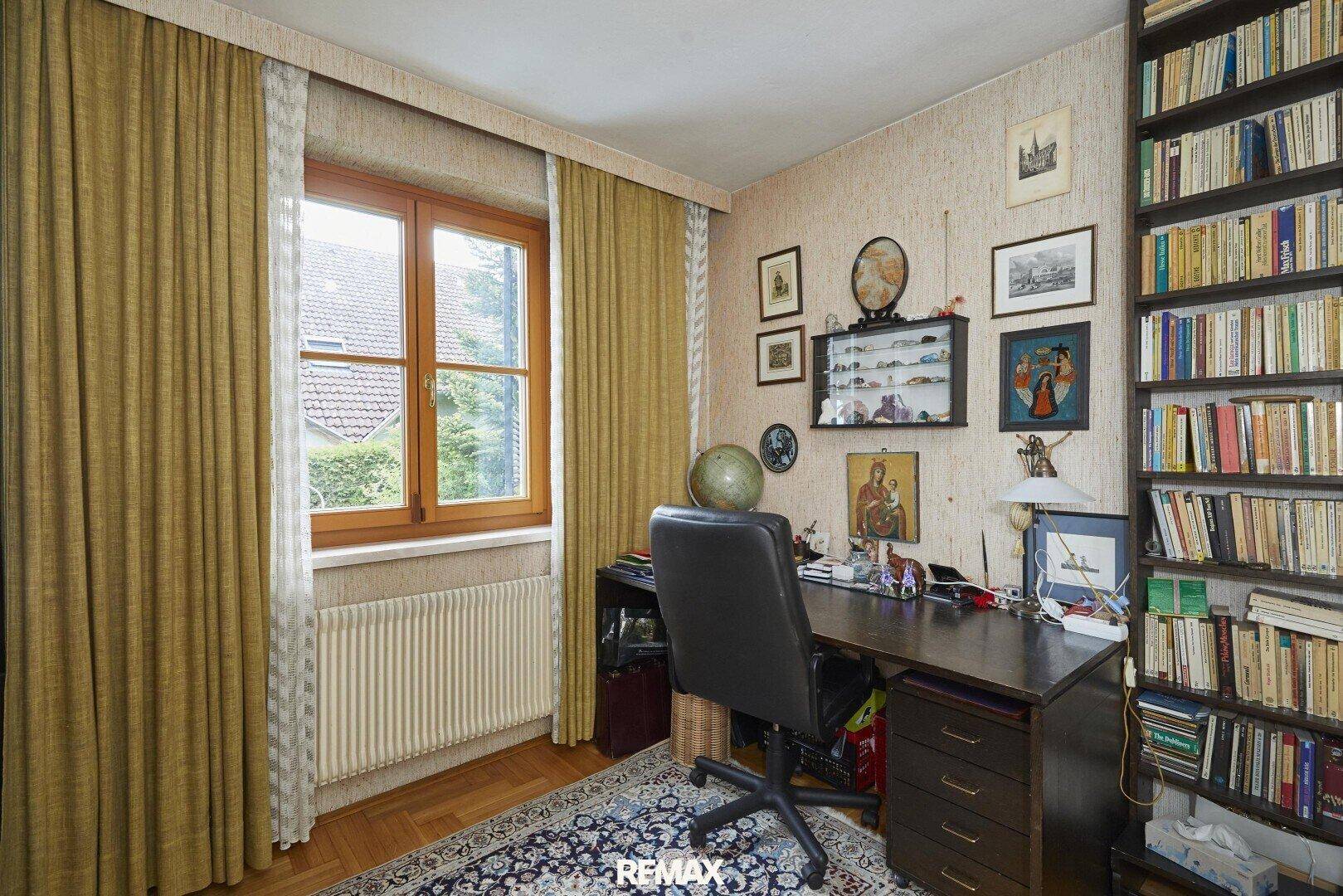 Arbeitszimmer mit Holzboden, großem Schreibtisch und Bücherregal am Fenster mit Tageslicht.