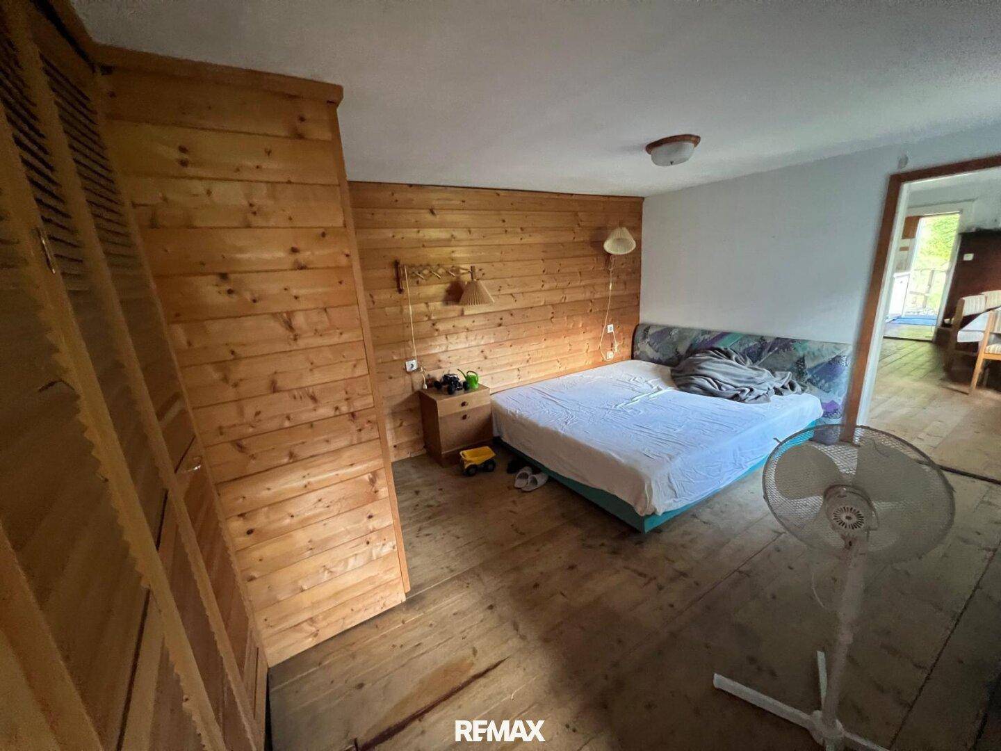 Rustikales Schlafzimmer mit Holzwänden und Dielenboden, ausgestattet mit einem Bett und Stehventilator.