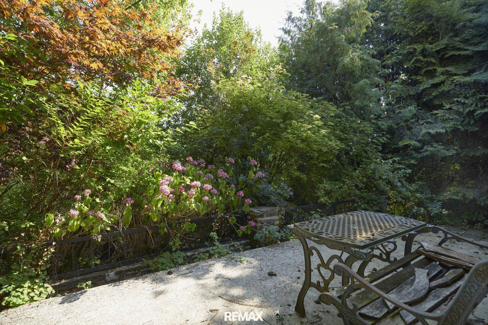 Gemütliche Terrasse mit schmiedeeisernen Gartenmöbeln, umgeben von üppiger Vegetation und Blumen.