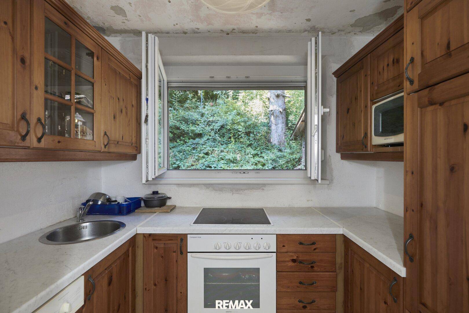 Rustikale Küche mit Holzschränken, weißer Arbeitsplatte und Blick ins Grüne durch das geöffnete Fenster.