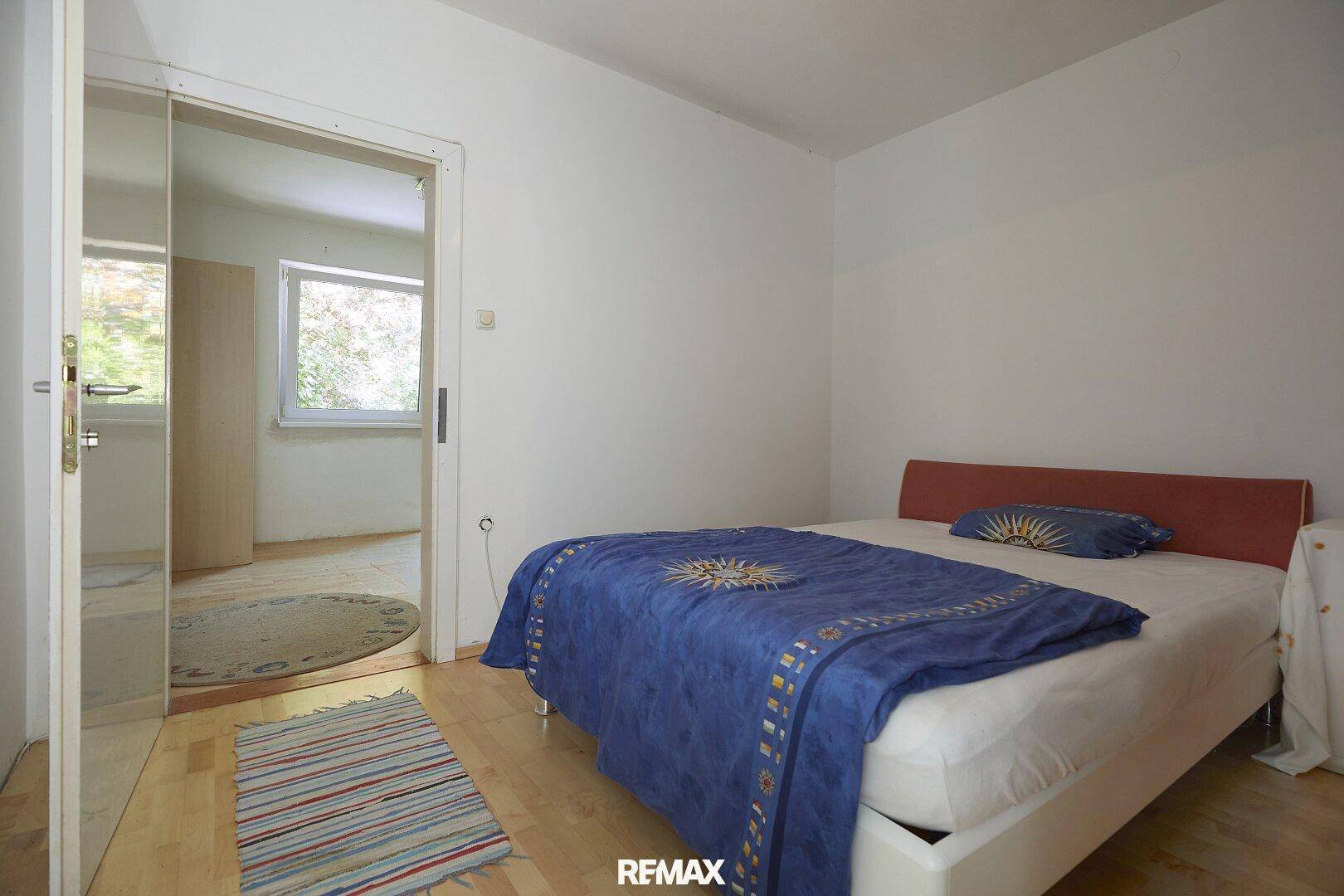 Helles Schlafzimmer mit Holzboden und einem blauen Bett, mit Zugang zu einem weiteren Raum.