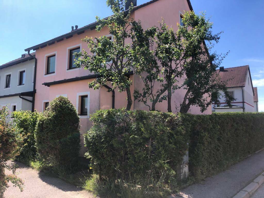 Seitenansicht des rosa Hauses mit hohen Hecken und Bäumen entlang der Straße.