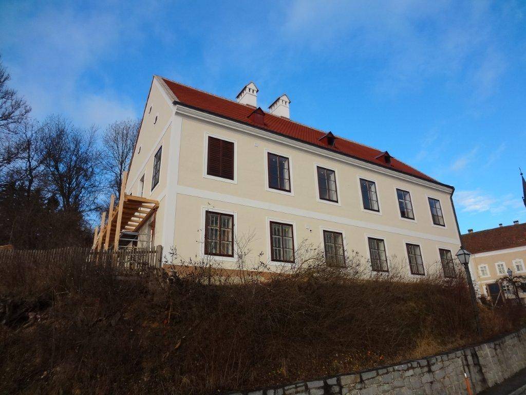 Historisches Gebäude mit heller Fassade, roten Dachziegeln und traditionellen Fenstern unter blauem Himmel.