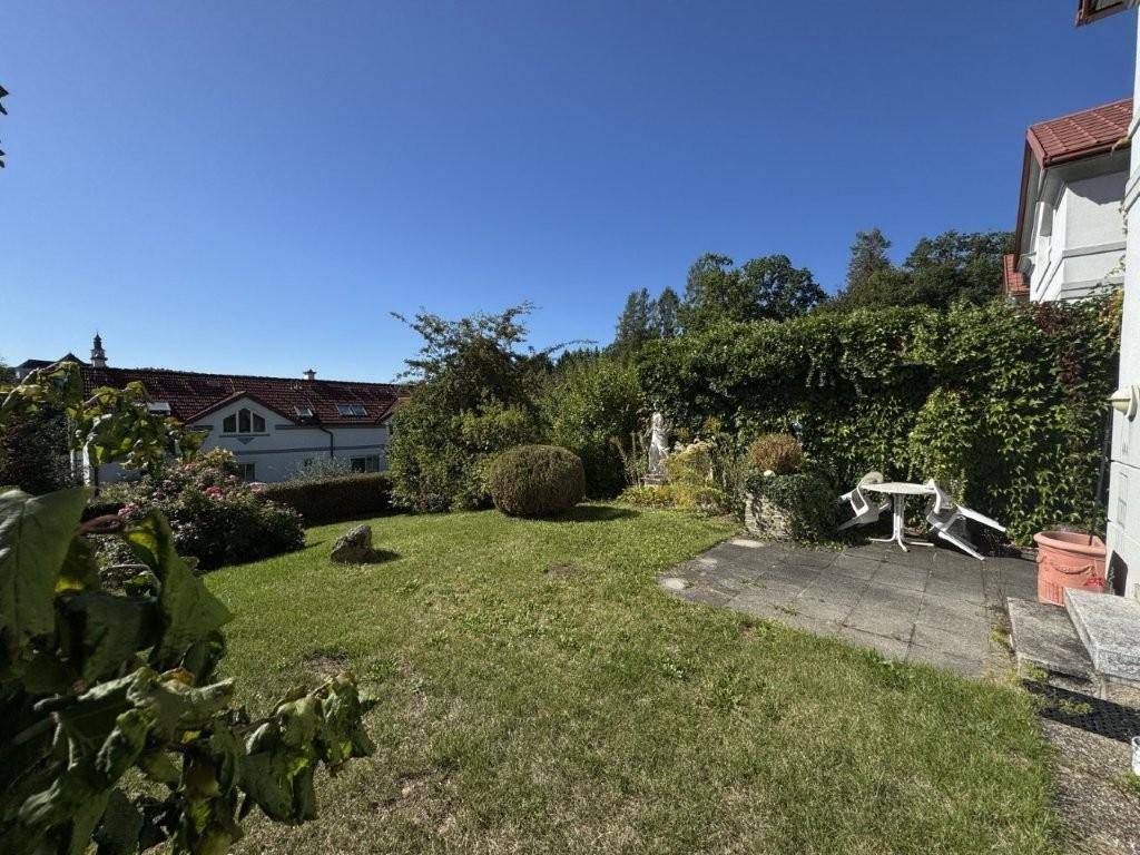 Gepflegter Gartenbereich mit Rasenfläche und kleiner Terrasse, umgeben von Hecken und Bäumen.