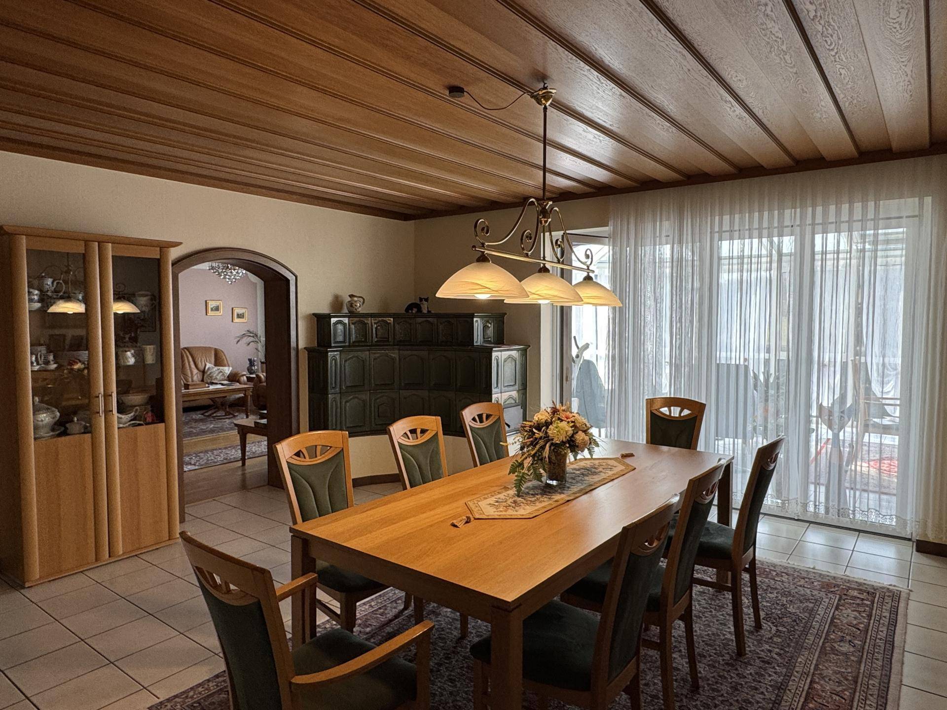Gemütlicher Essbereich mit Holztisch, Holzdecke und Zugang zur Terrasse, ergänzt durch einen Kachelofen.