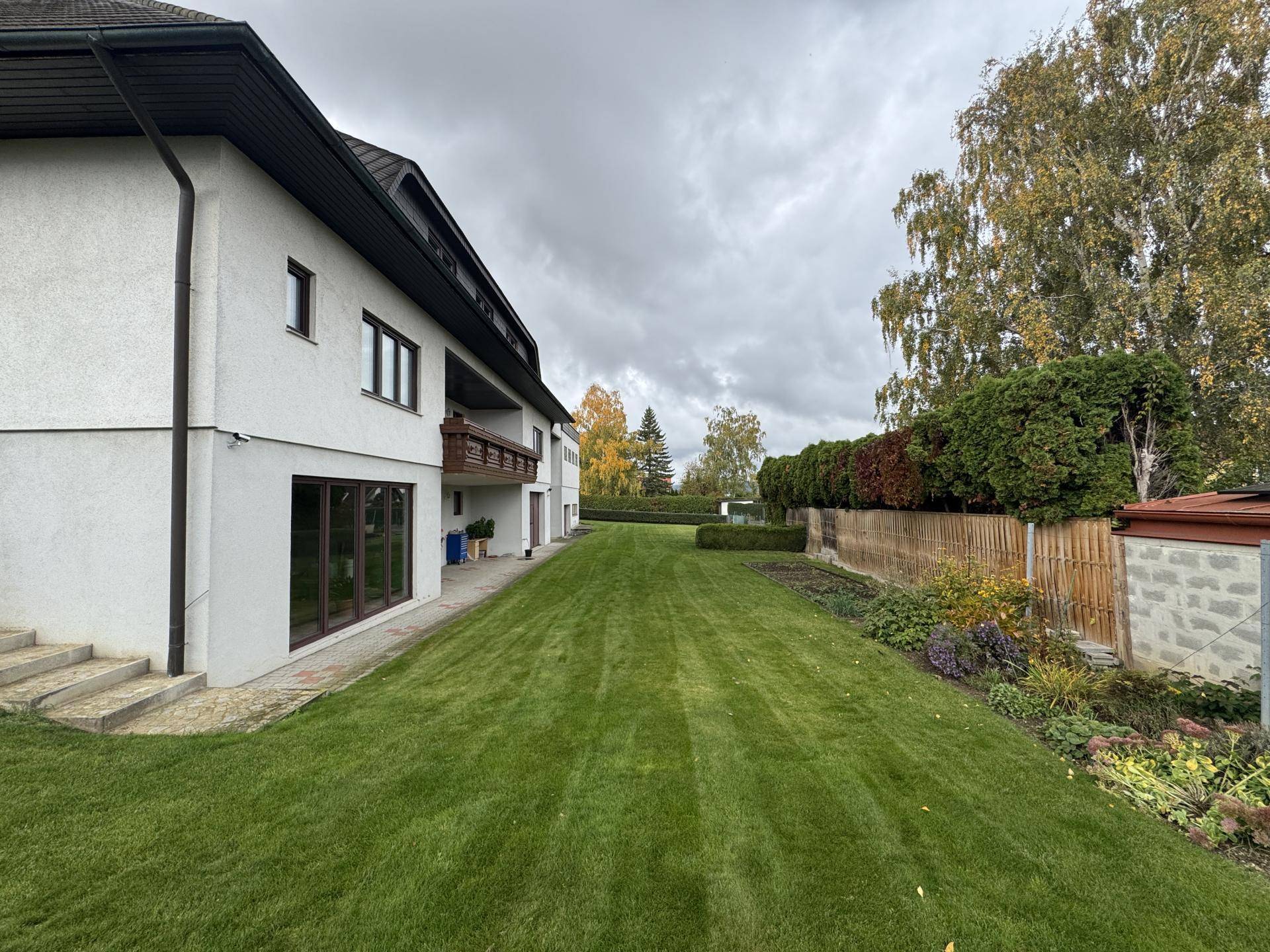 Seitenansicht des Hauses mit weitläufigem Rasenbereich und altem Baumbestand unter bewölktem Himmel.