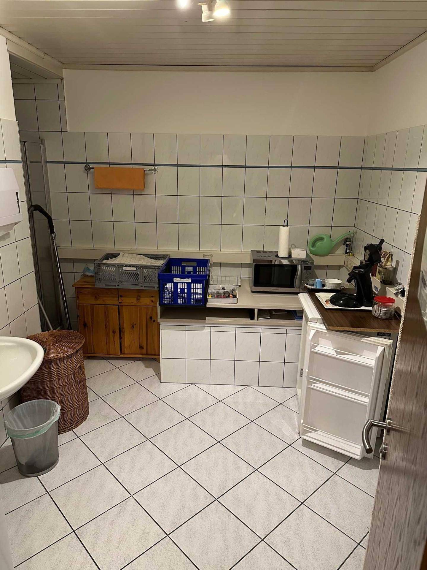 Kleine Küchenzeile mit weißen Fliesen, Spüle, Mikrowelle und Kühlschrank, funktional eingerichtet.