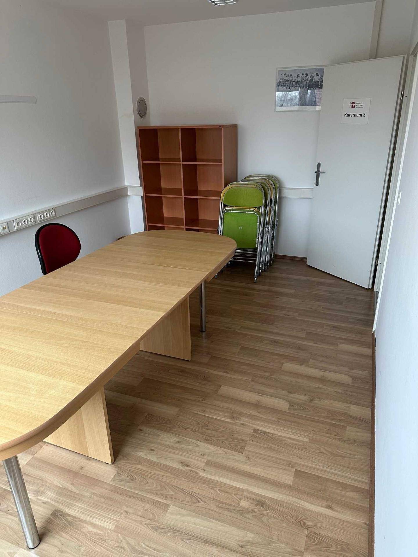 Einfacher Schulungsraum mit einem großen Tisch, Stühlen und einem Regal, ideal für kleine Gruppen.