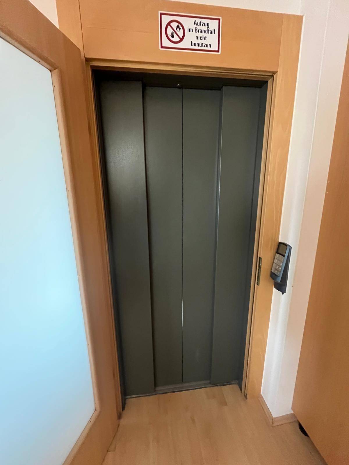Aufzug in einem Flur mit Holzverkleidung und einem Hinweisschild, bietet Zugang zu den Etagen.