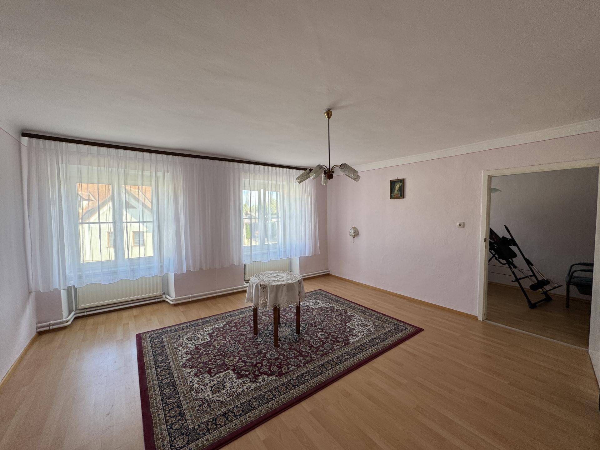 Helles Wohnzimmer mit Holzboden, Teppich und einem kleinen Tisch vor den Fenstern.