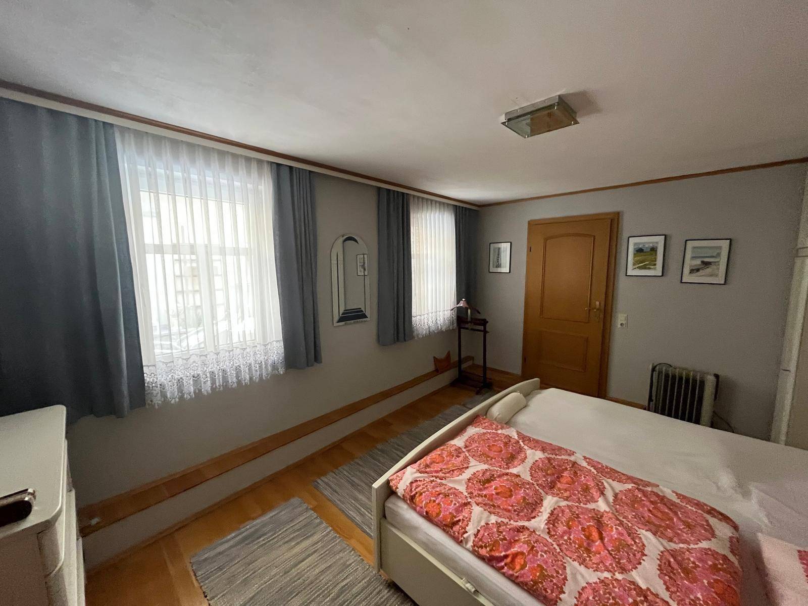 Schlafzimmer mit Holzboden, zwei Fenstern und einem Bett mit gemusterter Bettwäsche.