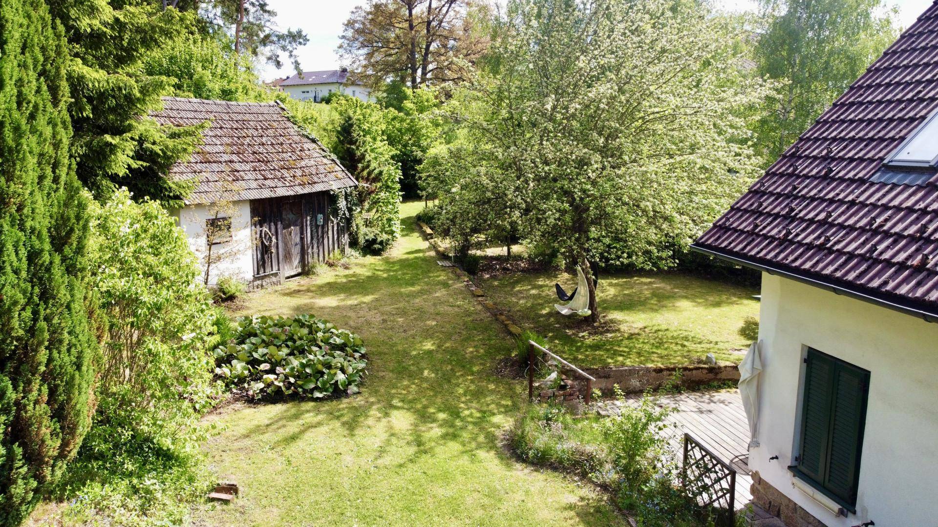 Weitläufiger grüner Garten mit altem Baumbestand und einem kleinen Nebengebäude im Hintergrund.