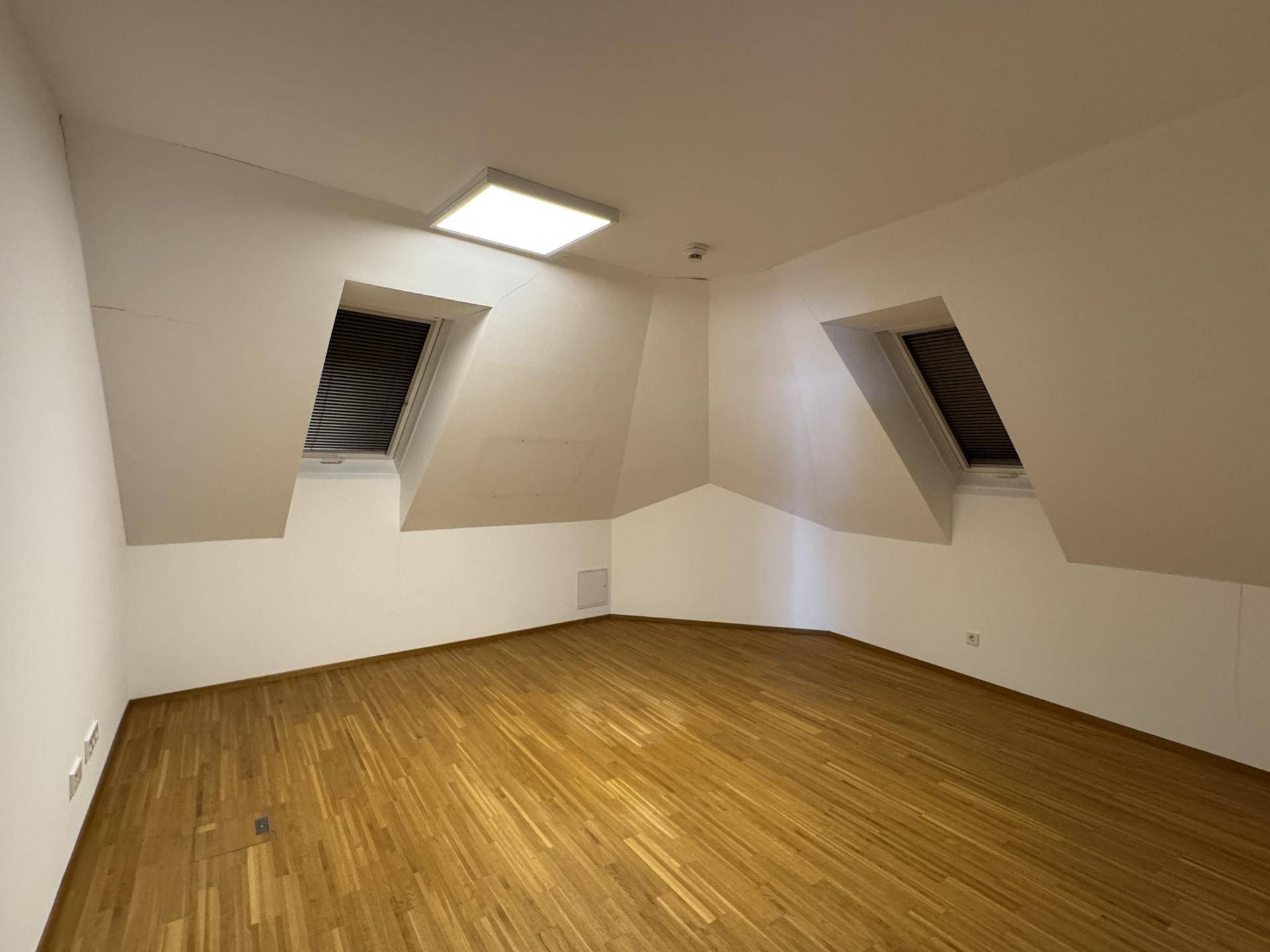 Leeres Zimmer mit Holzboden, Dachschrägen und zwei Dachfenstern, gut beleuchtet.