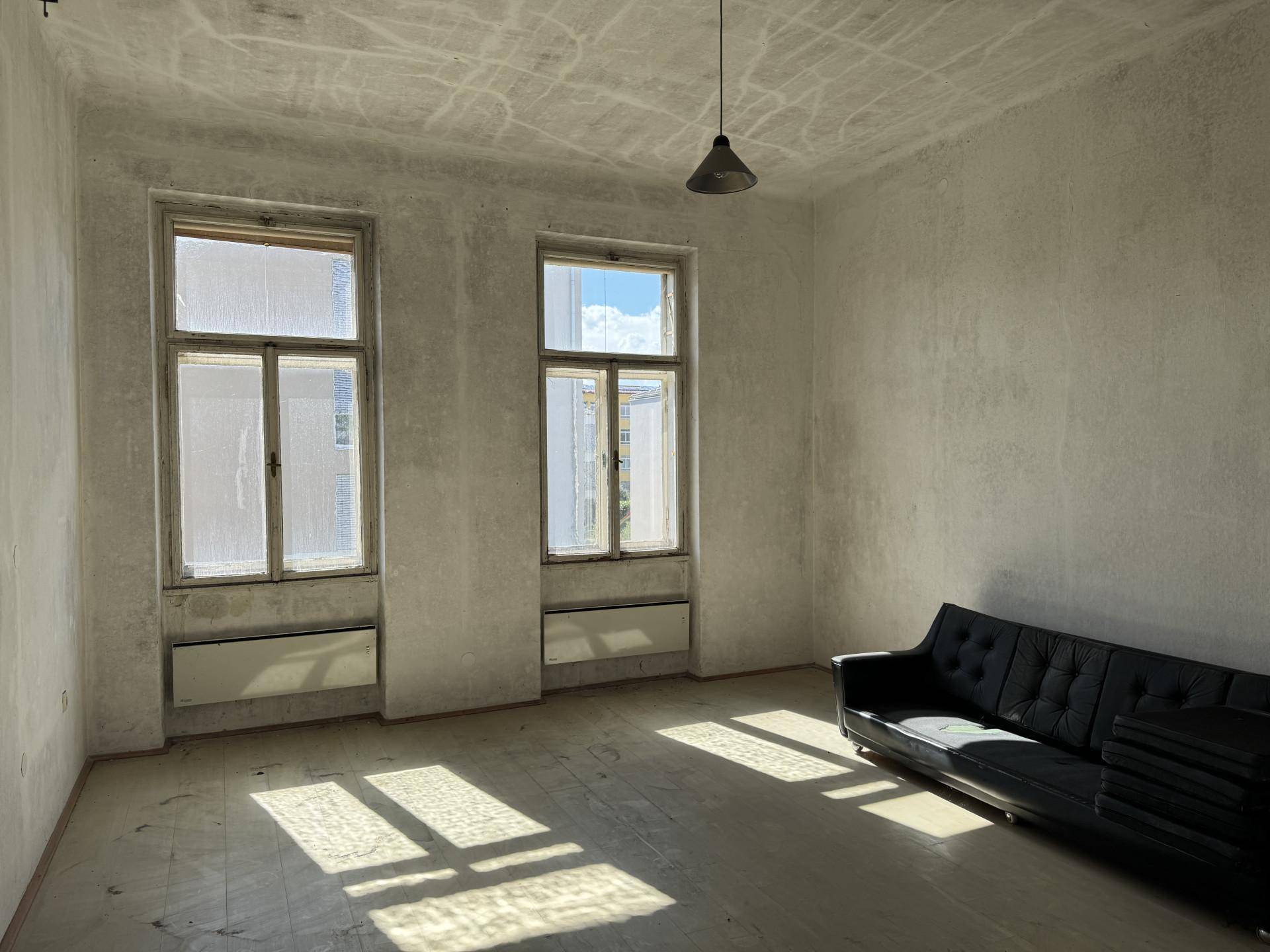 Helles Zimmer mit Holzboden, hohen Fenstern und einem schwarzen Sofa.