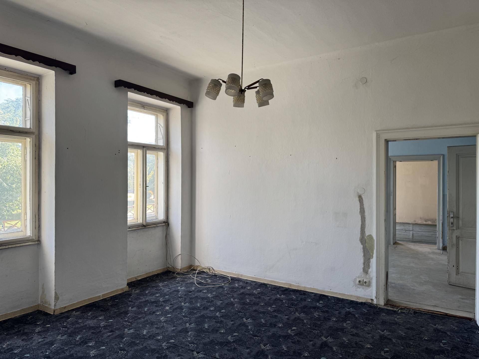 Geräumiges Zimmer mit dunklem Teppichboden, hohen Fenstern und einfacher Deckenleuchte.