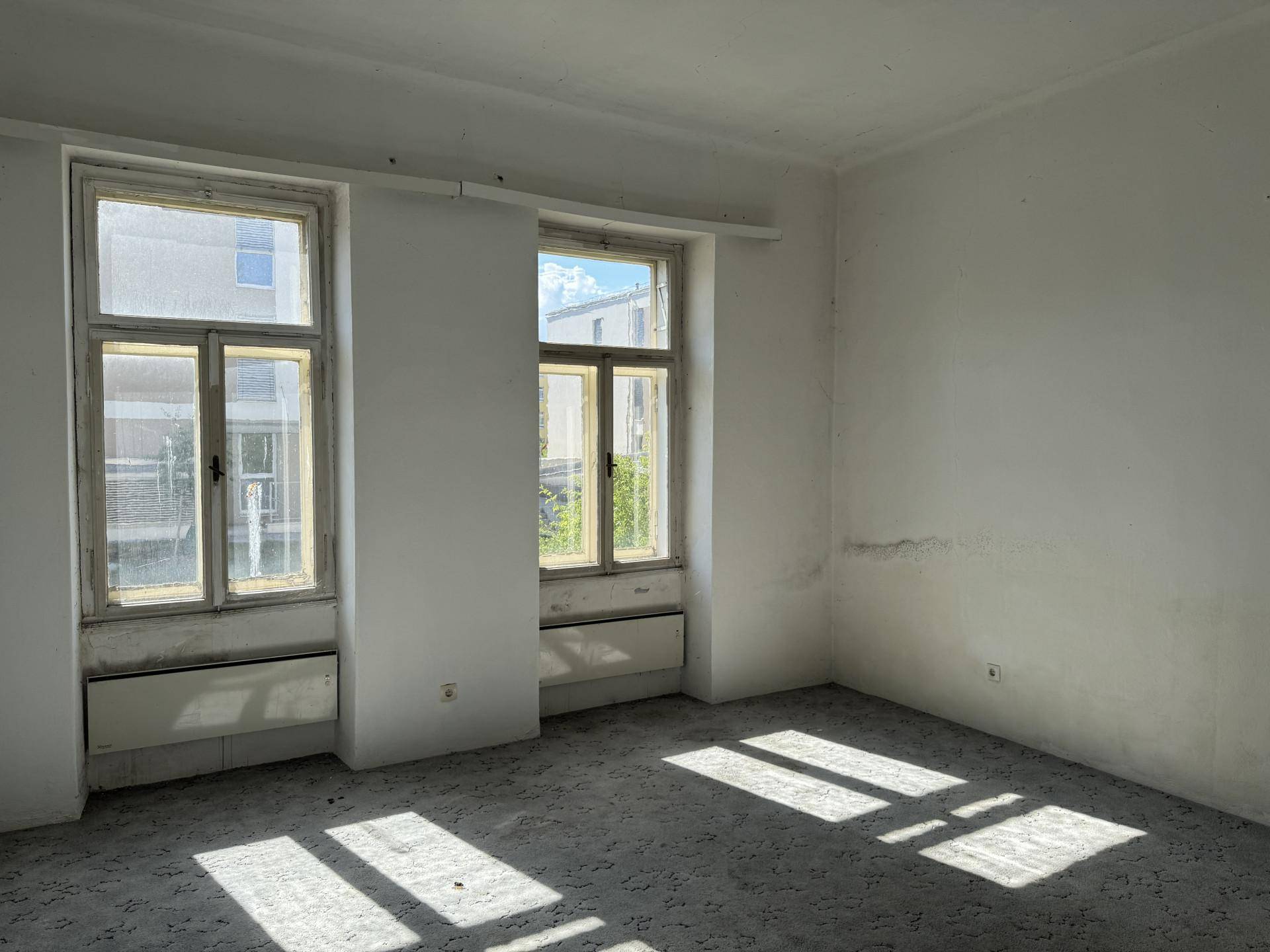 Helles Zimmer mit Teppichboden, hohen Fenstern und sichtbaren Gebrauchsspuren an den Wänden.