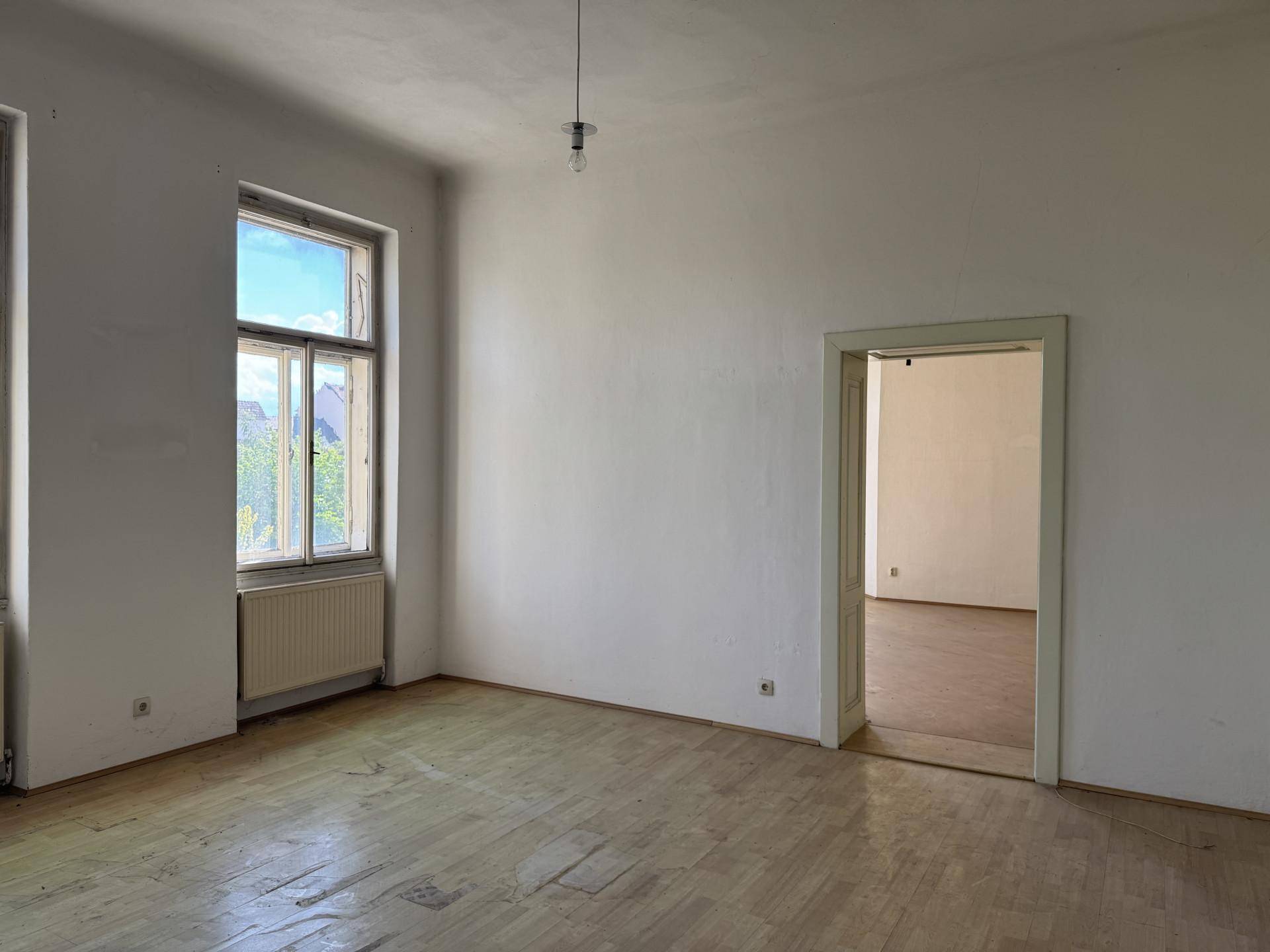 Geräumiges Zimmer mit Holzboden, hohen Fenstern und Zugang zu einem weiteren Raum.