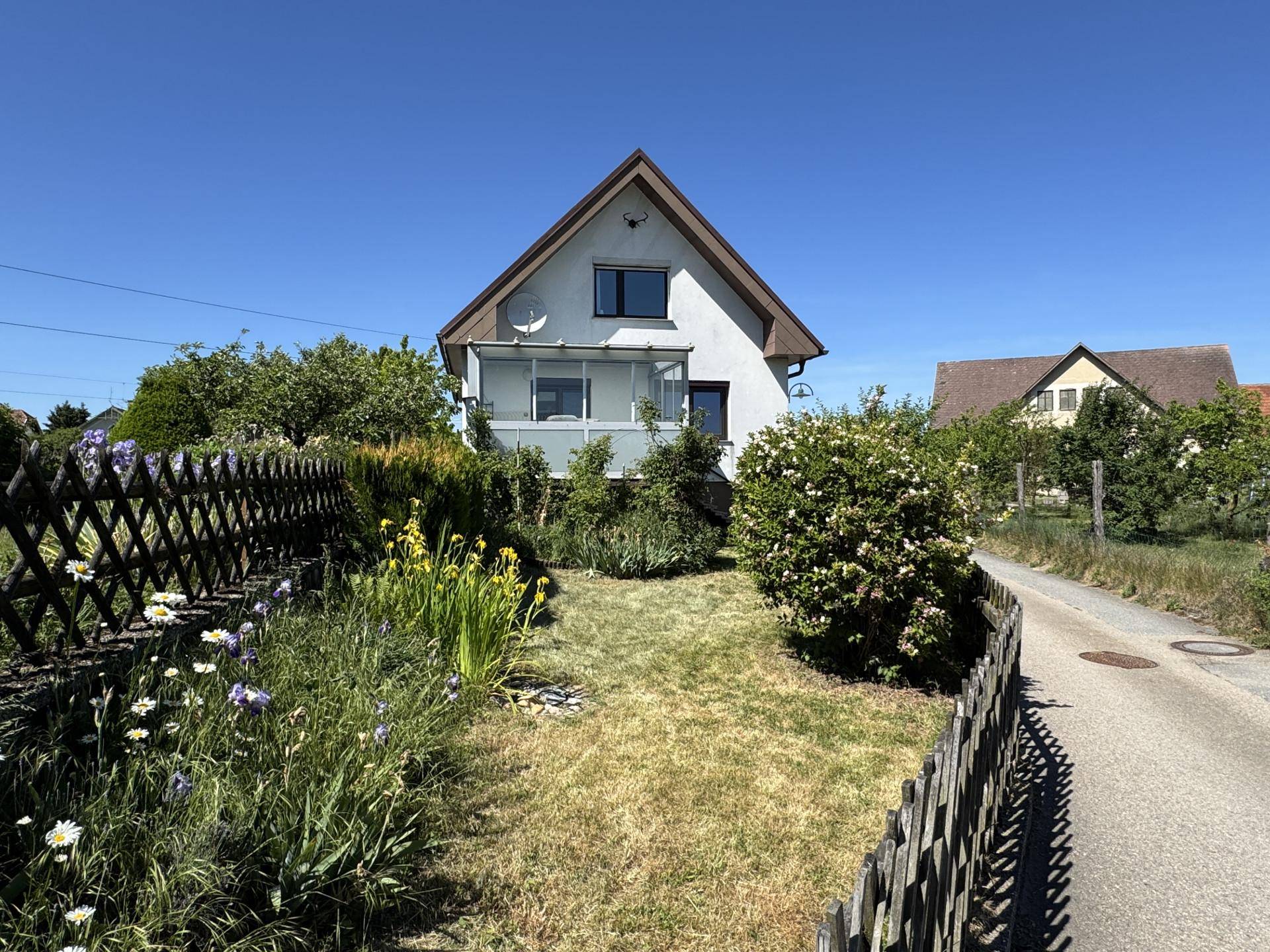 Sonniger Vorgarten mit Blumenbeeten und Blick auf das Einfamilienhaus mit verglastem Balkon.
