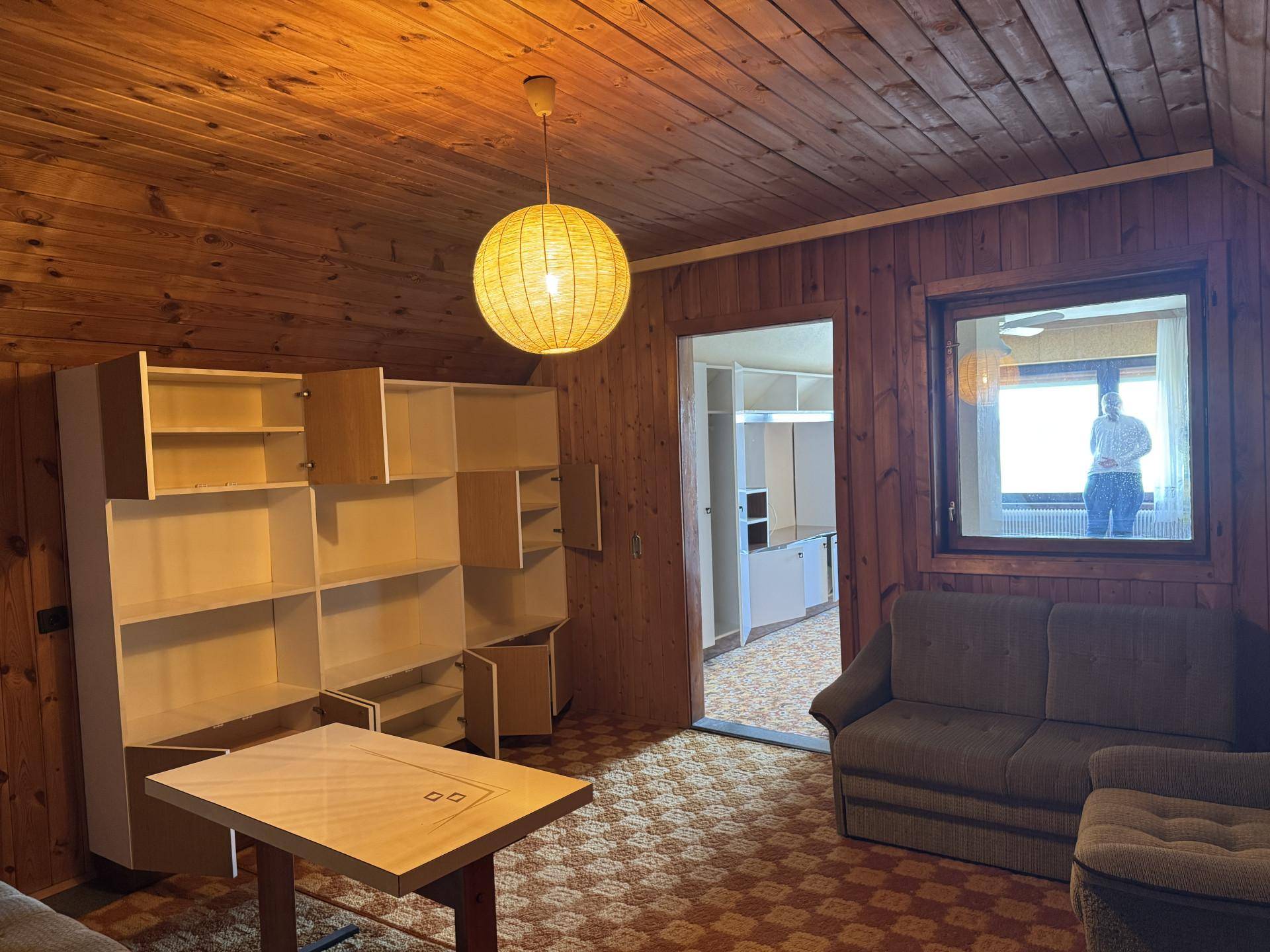 Schlafzimmer mit Holzvertäfelung, Teppichboden und einem Fenster mit Blick in einen weiteren Raum.