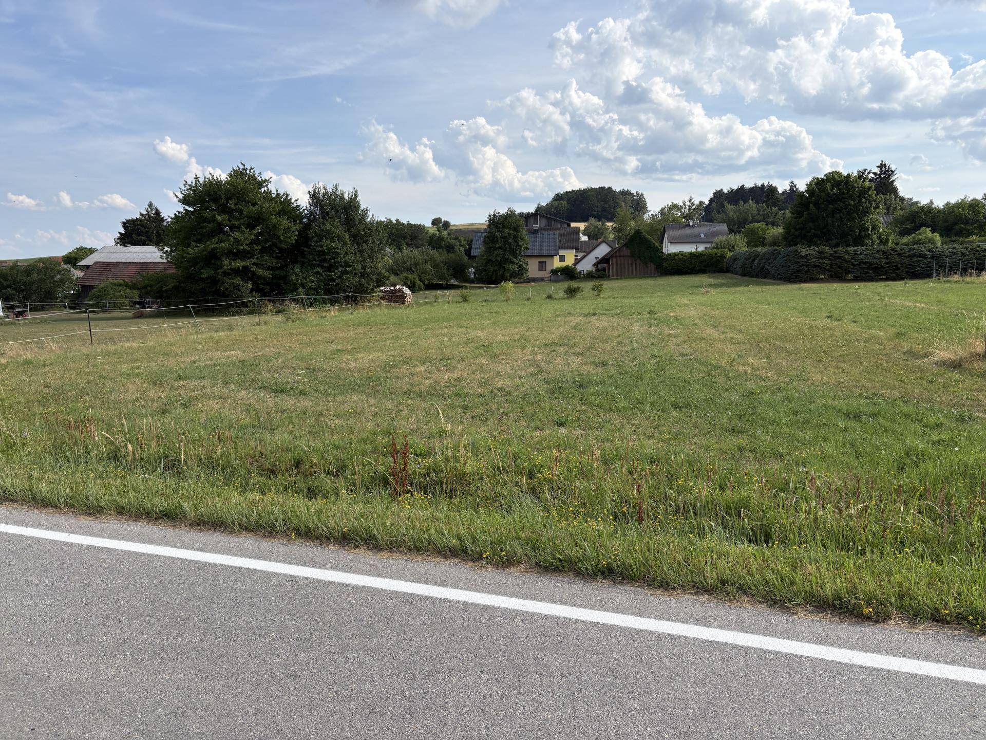 Blick von der Straße auf eine weitläufige Grünfläche mit ländlicher Umgebung und Dorf im Hintergrund.