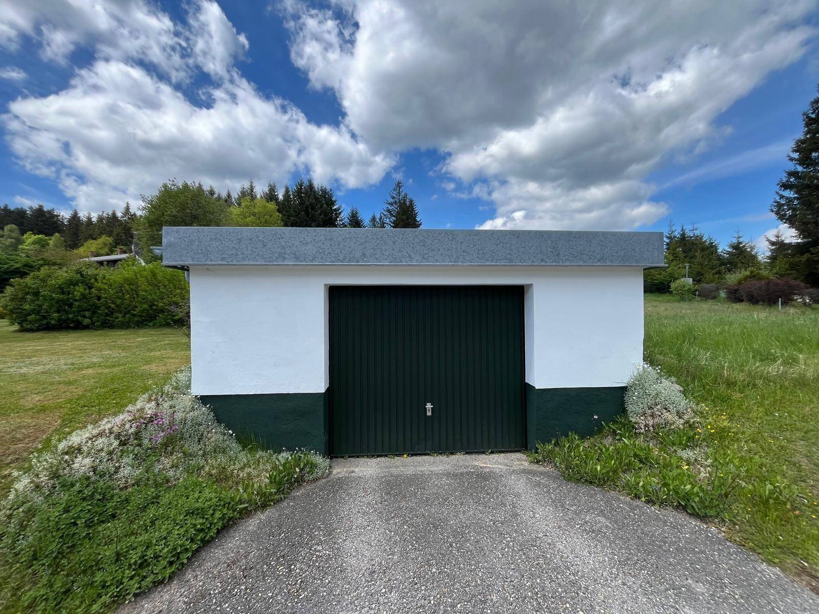 Einzelne Garage mit dunklem Tor und Zufahrt, umgeben von Gras und Büschen.