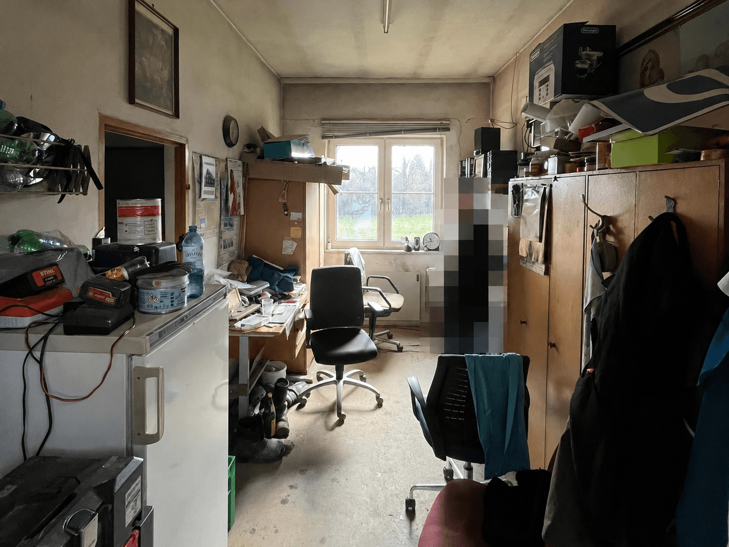 Überfülltes Büro oder Mehrzweckraum mit Schreibtisch, Stühlen, Kühlschrank und einem Fenster mit Blick ins Grüne.