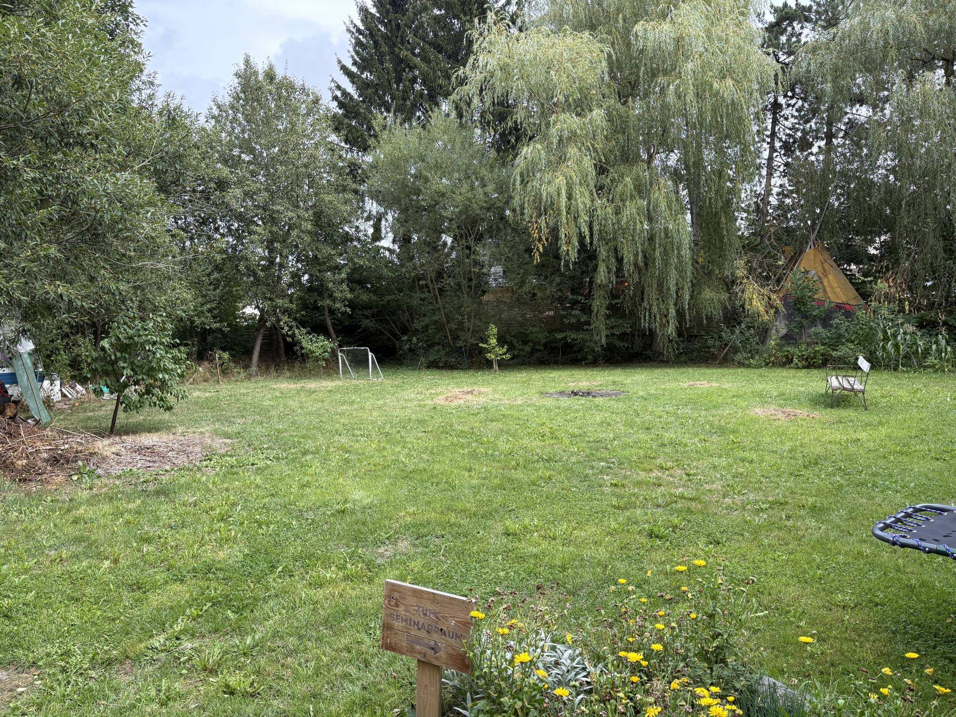 Großer, grüner Garten mit Rasenfläche, Bäumen und einer Feuerstelle im Freien.