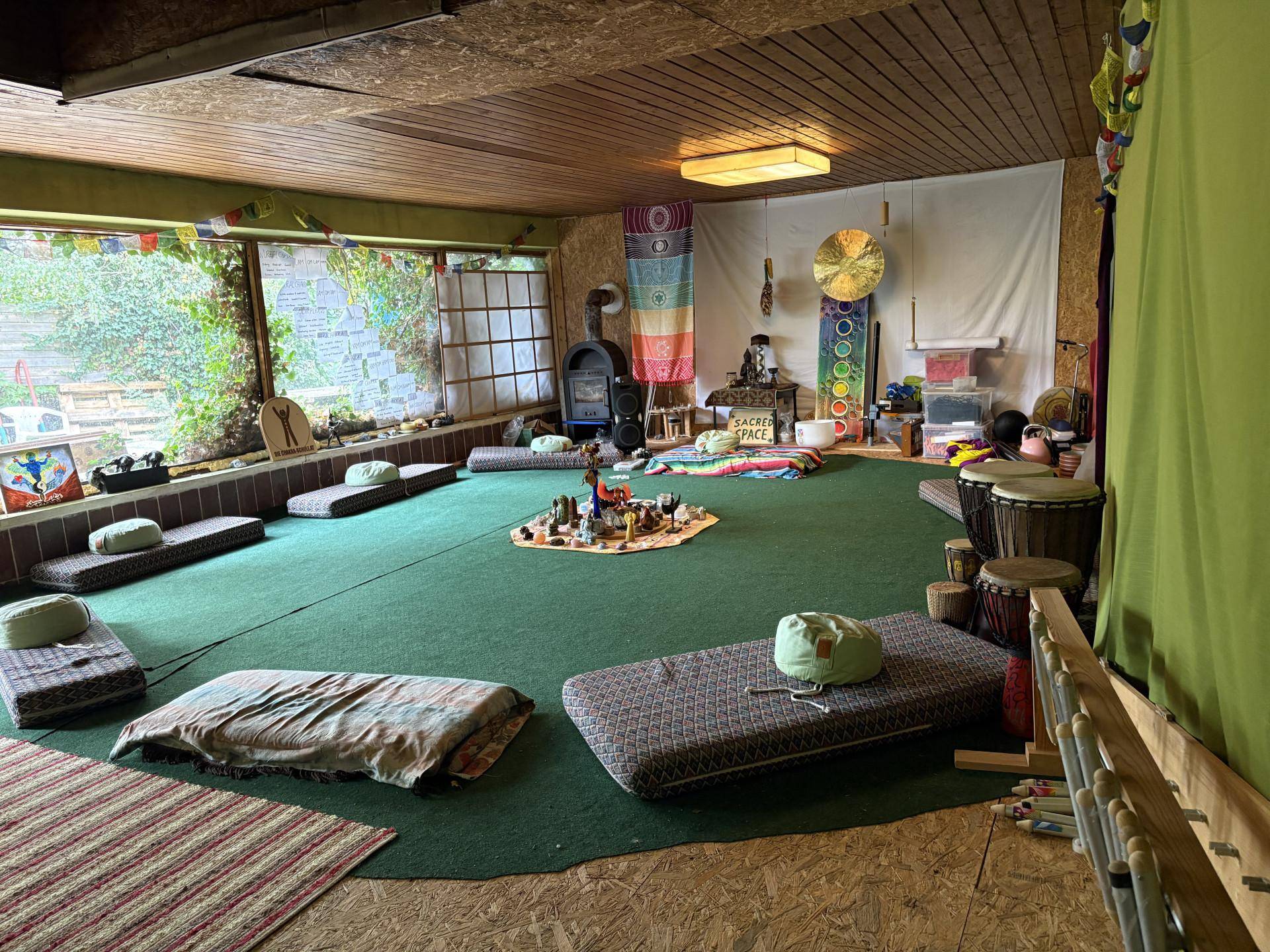 Großer Gemeinschaftsraum mit grünem Teppich, Holzdecke und Sitzkissen für Meditation.