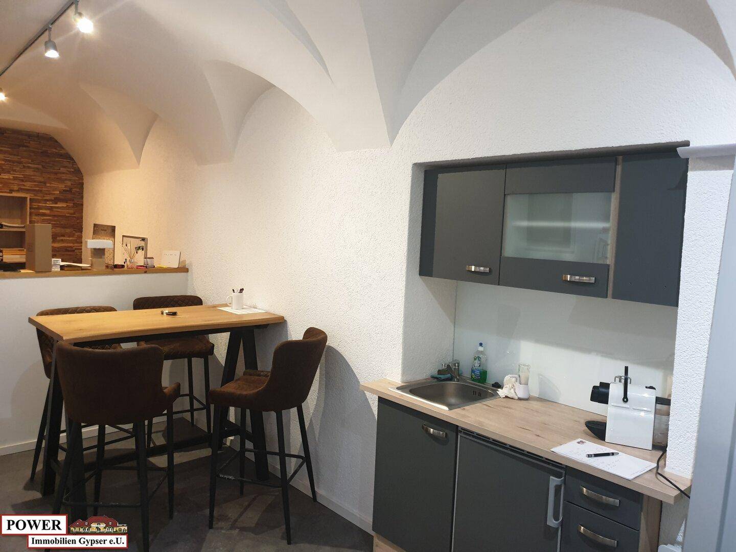 Kleine, moderne Küchenzeile mit grauen Unterschränken, Holz-Arbeitsplatte und Spülbecken in einem Geschäftsraum. Die Gewölbedecke ist auch hier sichtbar.
