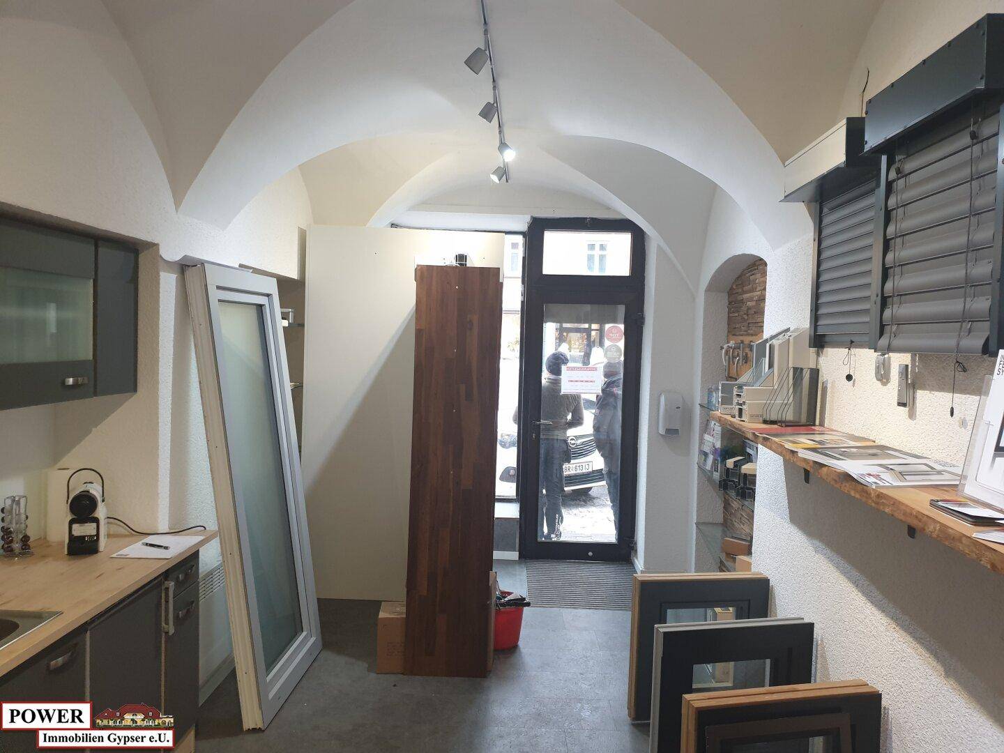 Eingangsbereich eines Geschäftsraums mit Glastür, Gewölbedecke und Ausstellungsregalen. Die Fenster lassen viel natürliches Licht herein.