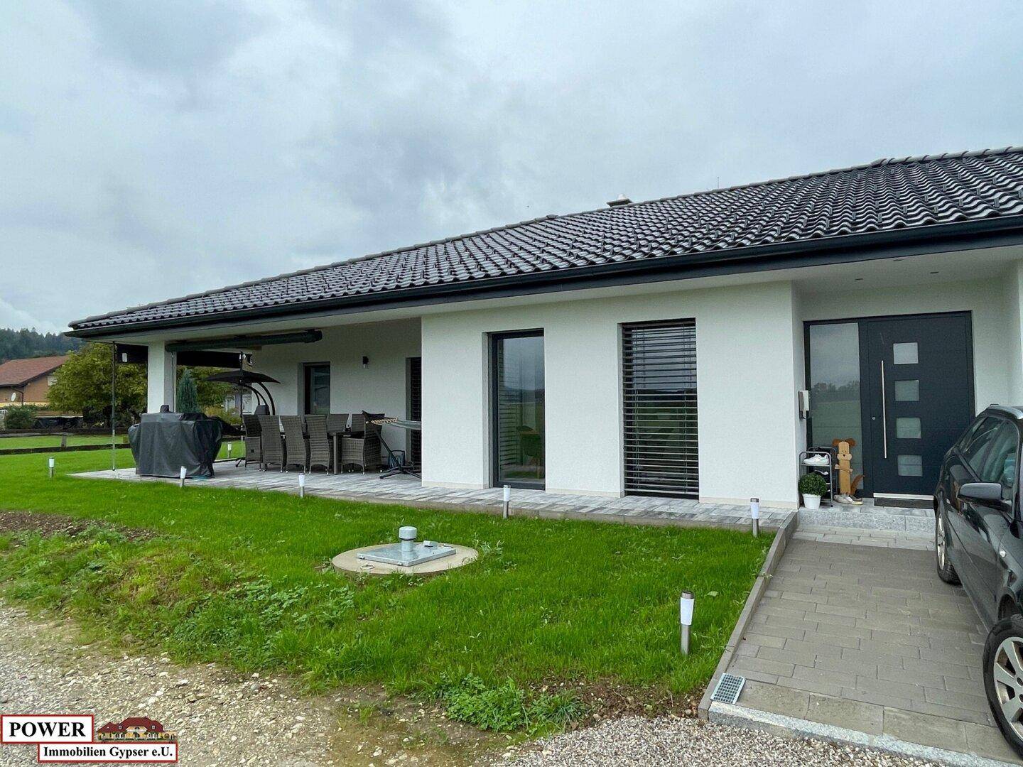 Überdachte Terrasse mit Esstisch und Stühlen, umgeben von Rasen und Blick auf die Landschaft.