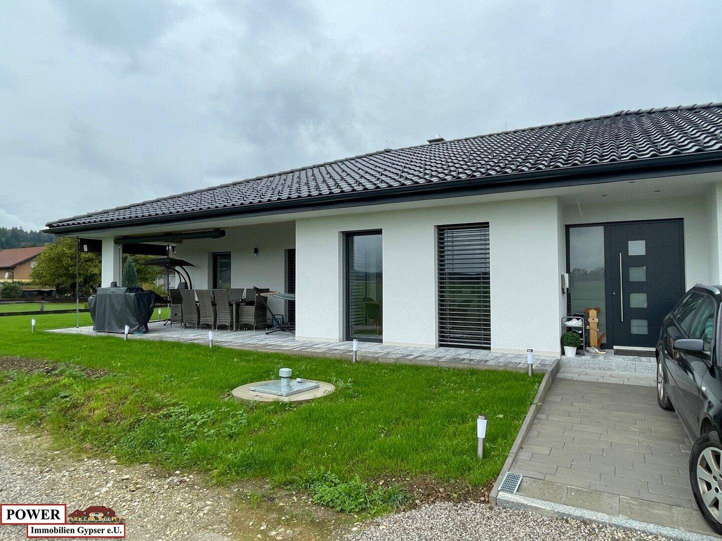Überdachte Terrasse mit Gartenmöbeln und Zugang zum Haus, umgeben von Rasen.