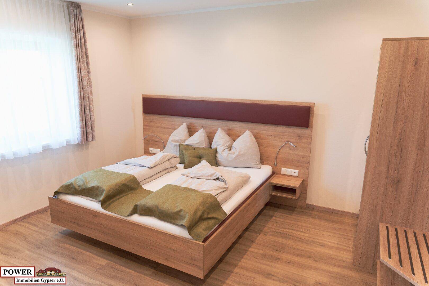 Helles Schlafzimmer mit Doppelbett, Holzmöbeln und Fenster mit Vorhängen.