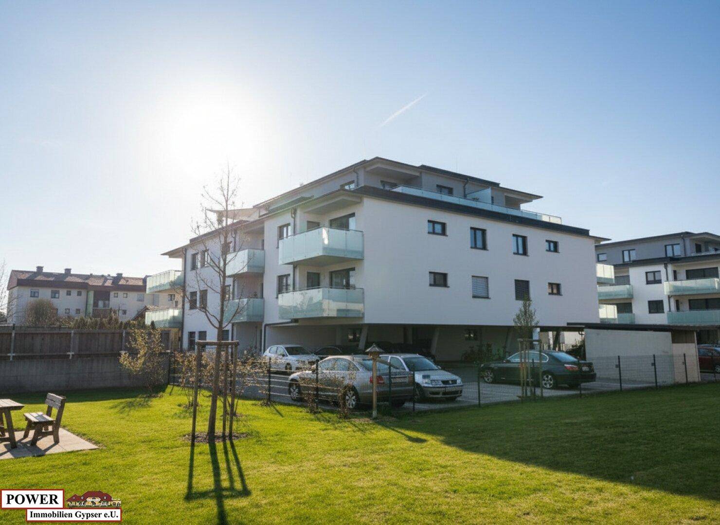 Modernes Mehrfamilienhaus mit Balkonen, Parkplätzen und Grünflächen unter strahlendem Himmel.