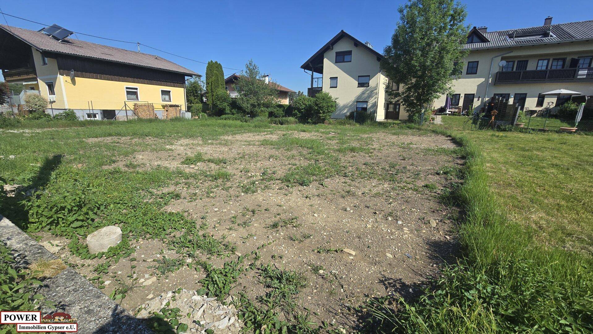 Offenes Baugrundstück mit leichter Vegetation, umgeben von Einfamilienhäusern in ländlicher Umgebung.