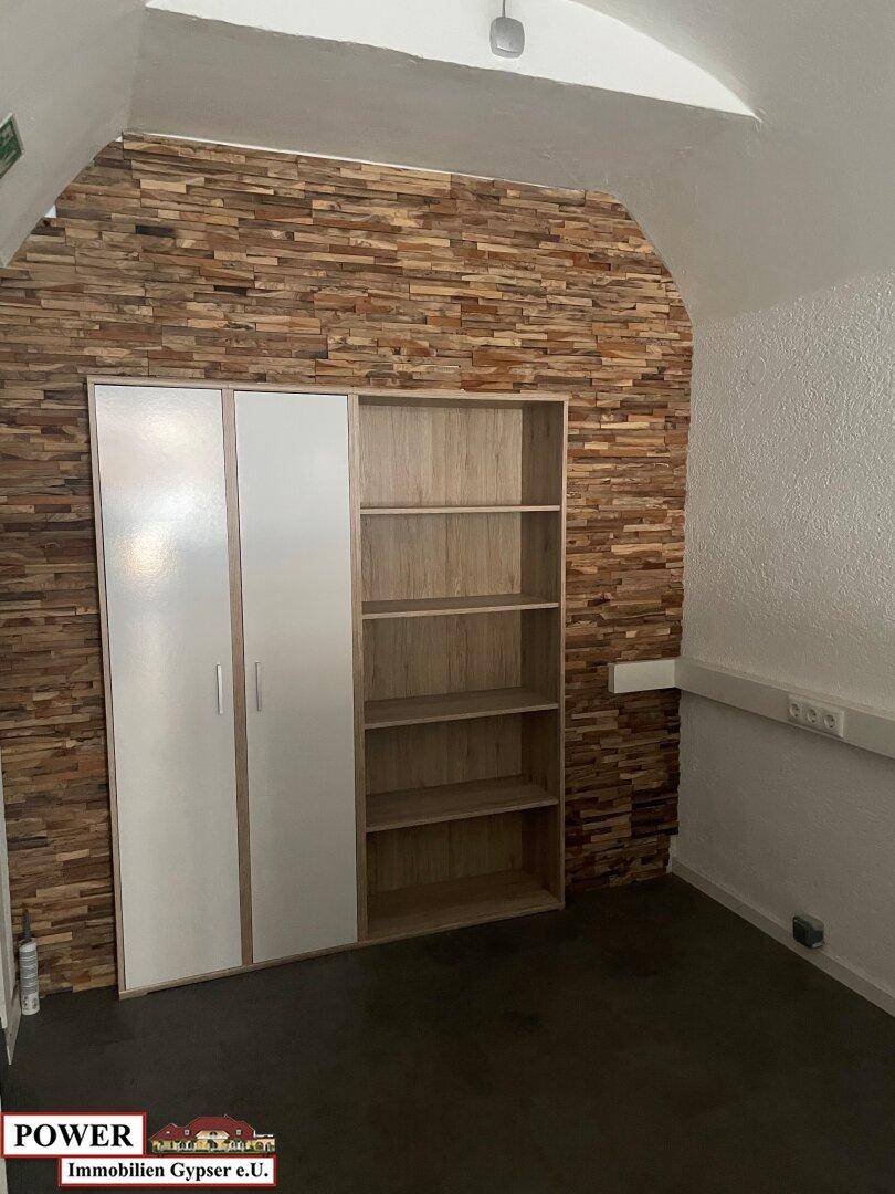 Bereich mit weißen Schränken und offenen Holzregalen vor einer auffälligen Holzwand in einem Geschäftsraum. Der Boden ist dunkel gehalten.