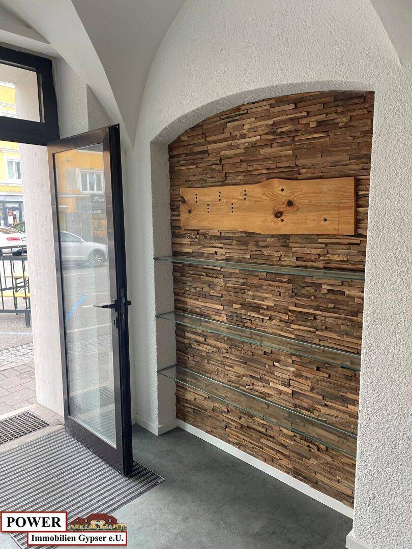Eingangsbereich mit offener Glastür, einer dekorativen Holzwand und integrierten Glasregalen. Die Gewölbedecke ist ein Blickfang.