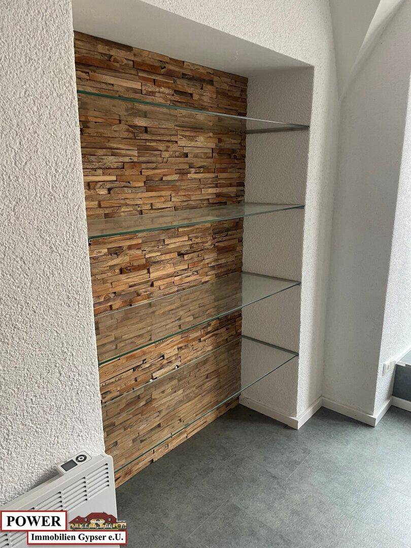 Nische mit dekorativer Holzwand und mehreren Glasregalen in einem Geschäftsraum. Ein Heizkörper ist unter den Regalen angebracht.