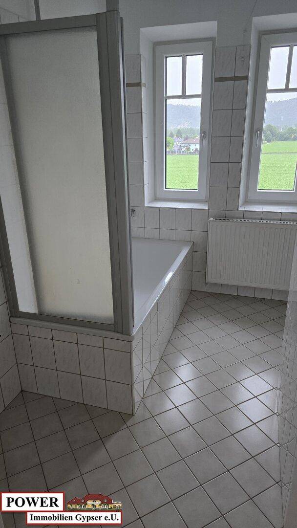 Helles Badezimmer mit Badewanne, Duschabtrennung und zwei Fenstern mit Blick ins Grüne.