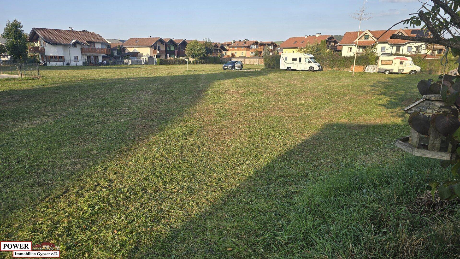 Weitläufige Wiese mit Blick auf eine Wohnsiedlung und geparkte Fahrzeuge in der Ferne.