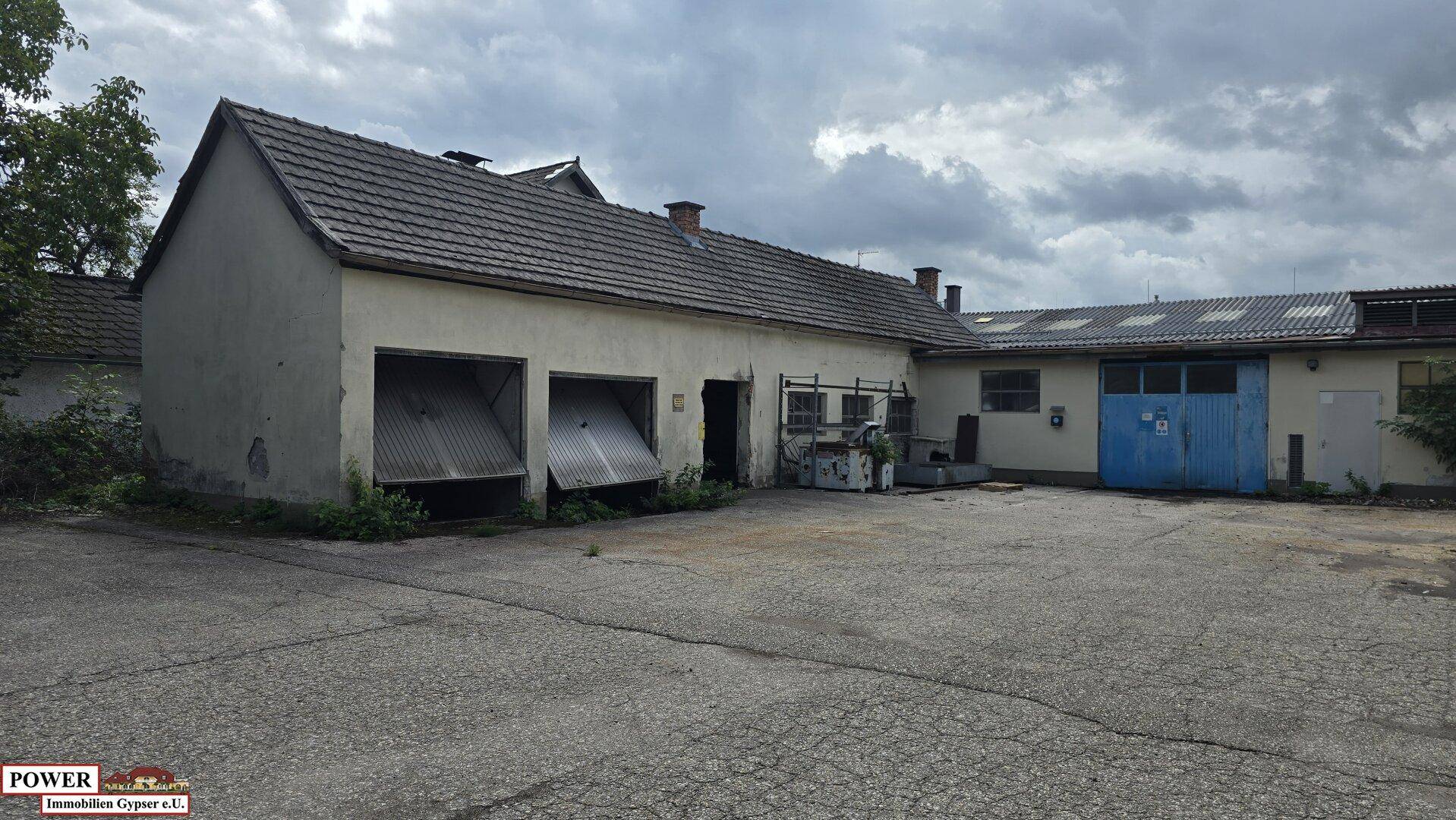 Gebäudeteil mit zwei Garagentoren und einem großen, asphaltierten Vorplatz unter einem bewölkten Himmel.
