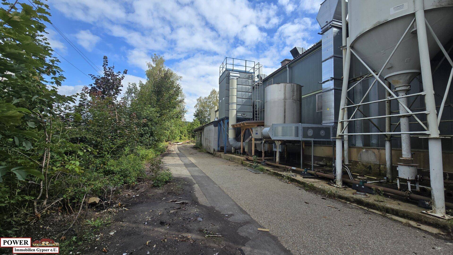 Seitenansicht eines Industriegebäudes mit großen Silos und Lüftungsanlagen, umgeben von Bäumen.