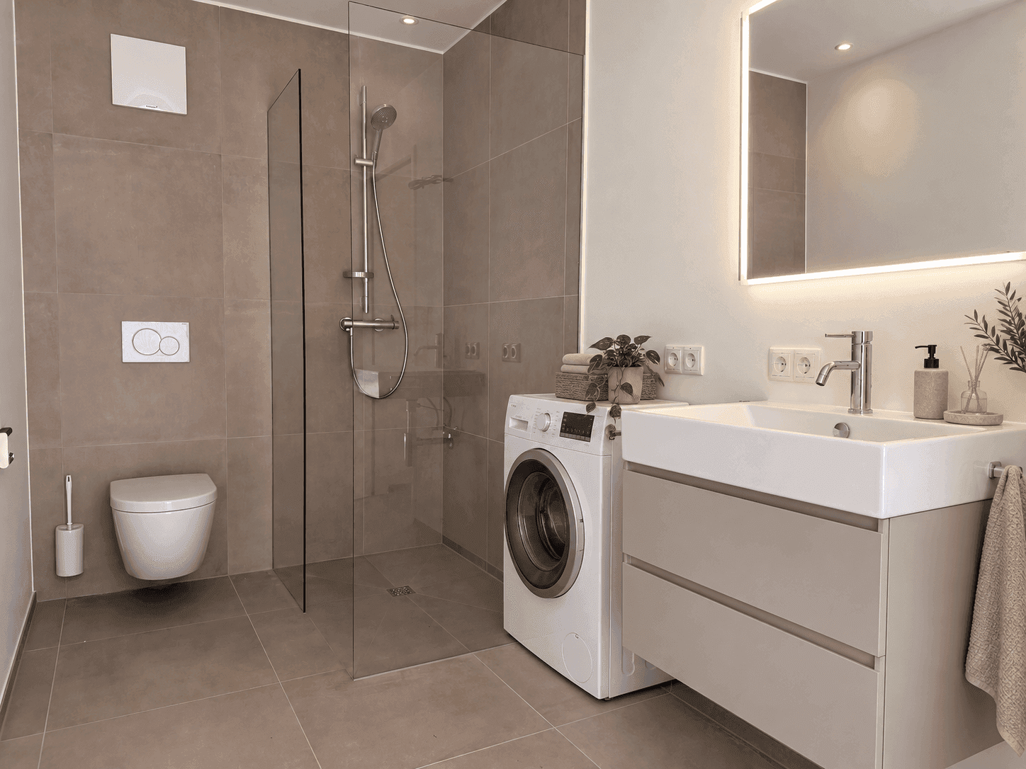 Modernes Badezimmer mit ebenerdiger Dusche, Waschmaschine und beleuchtetem Spiegel über dem Waschtisch.