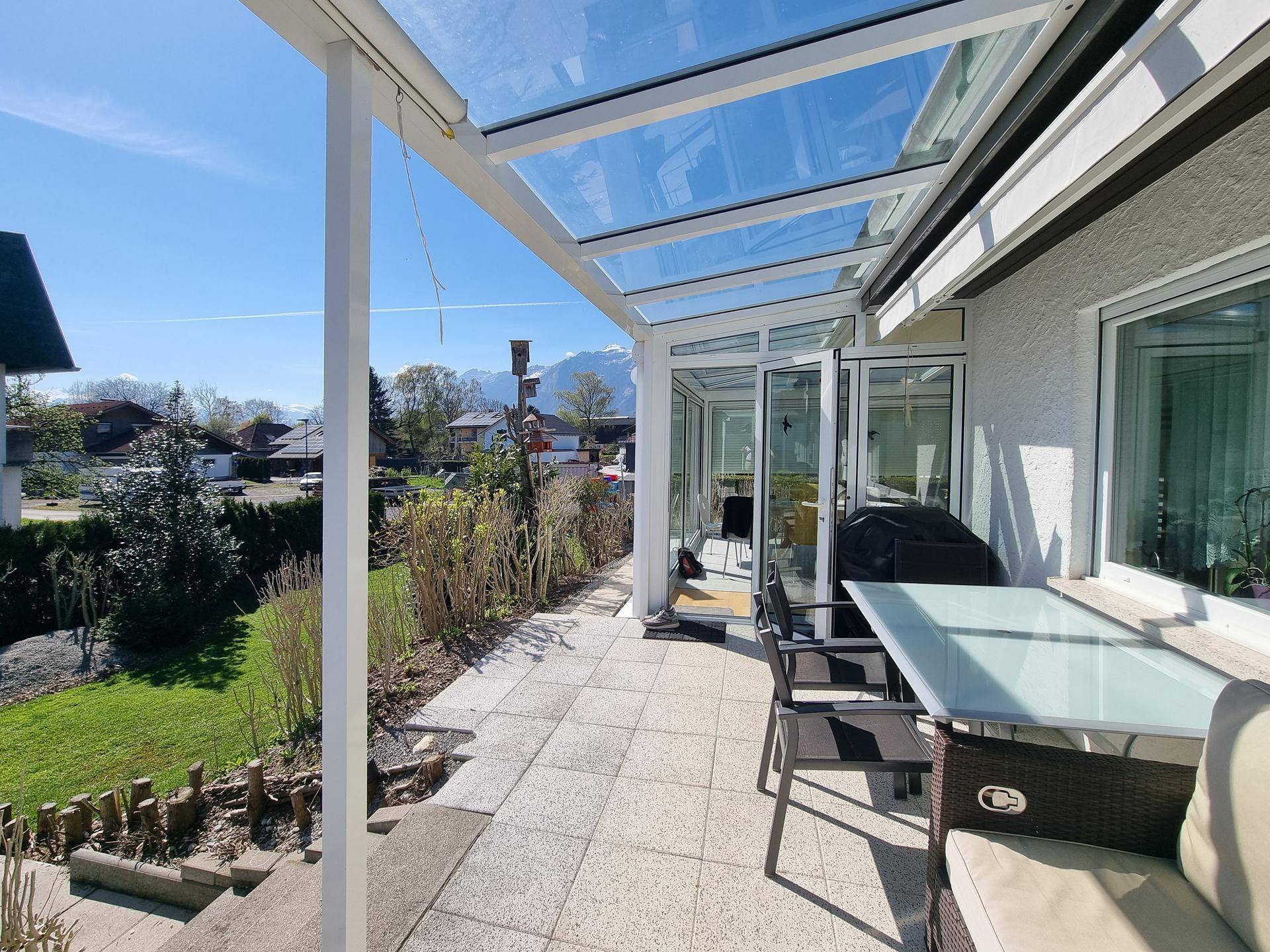Sonnige Terrasse mit Glasüberdachung, Gartenmöbeln und Blick auf die Berge.