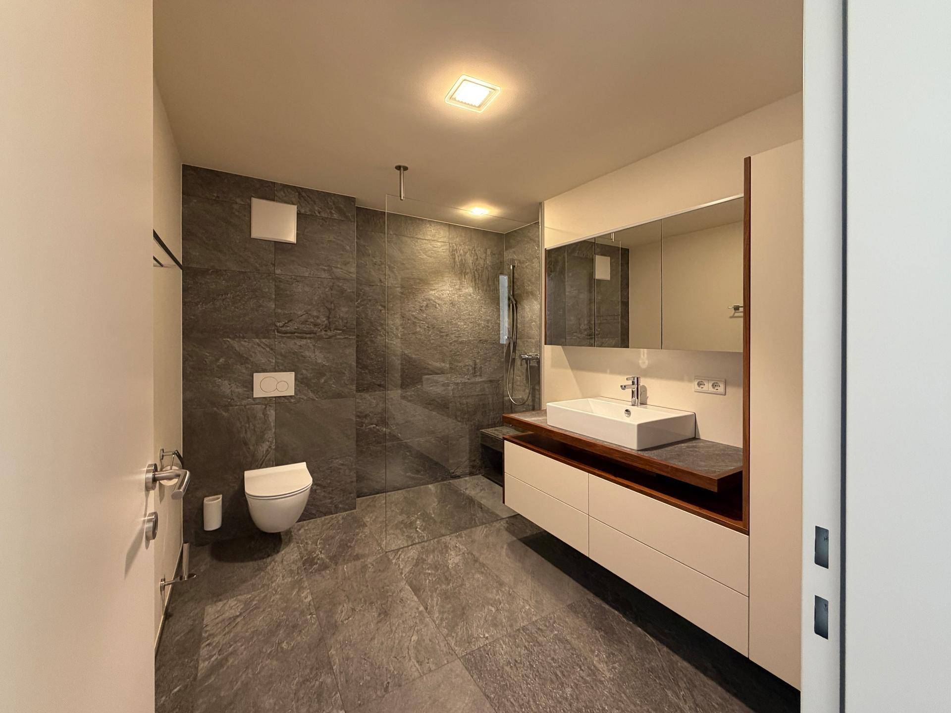 Modernes Badezimmer mit dunklen Fliesen, begehbarer Dusche und elegantem Waschtisch.