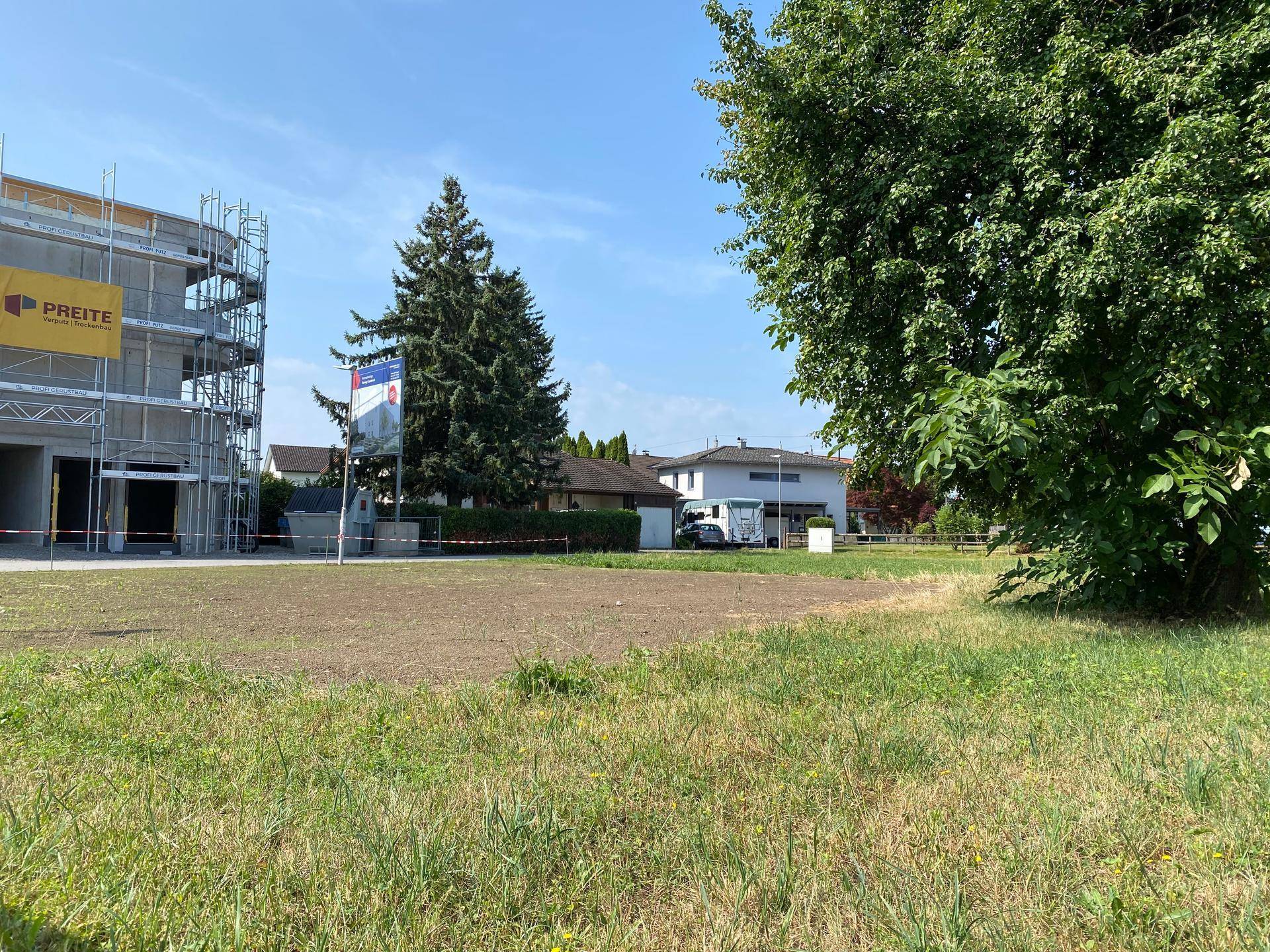 Blick auf ein unbebautes Grundstück mit einer Baustelle im Hintergrund und weiteren Wohnhäusern.