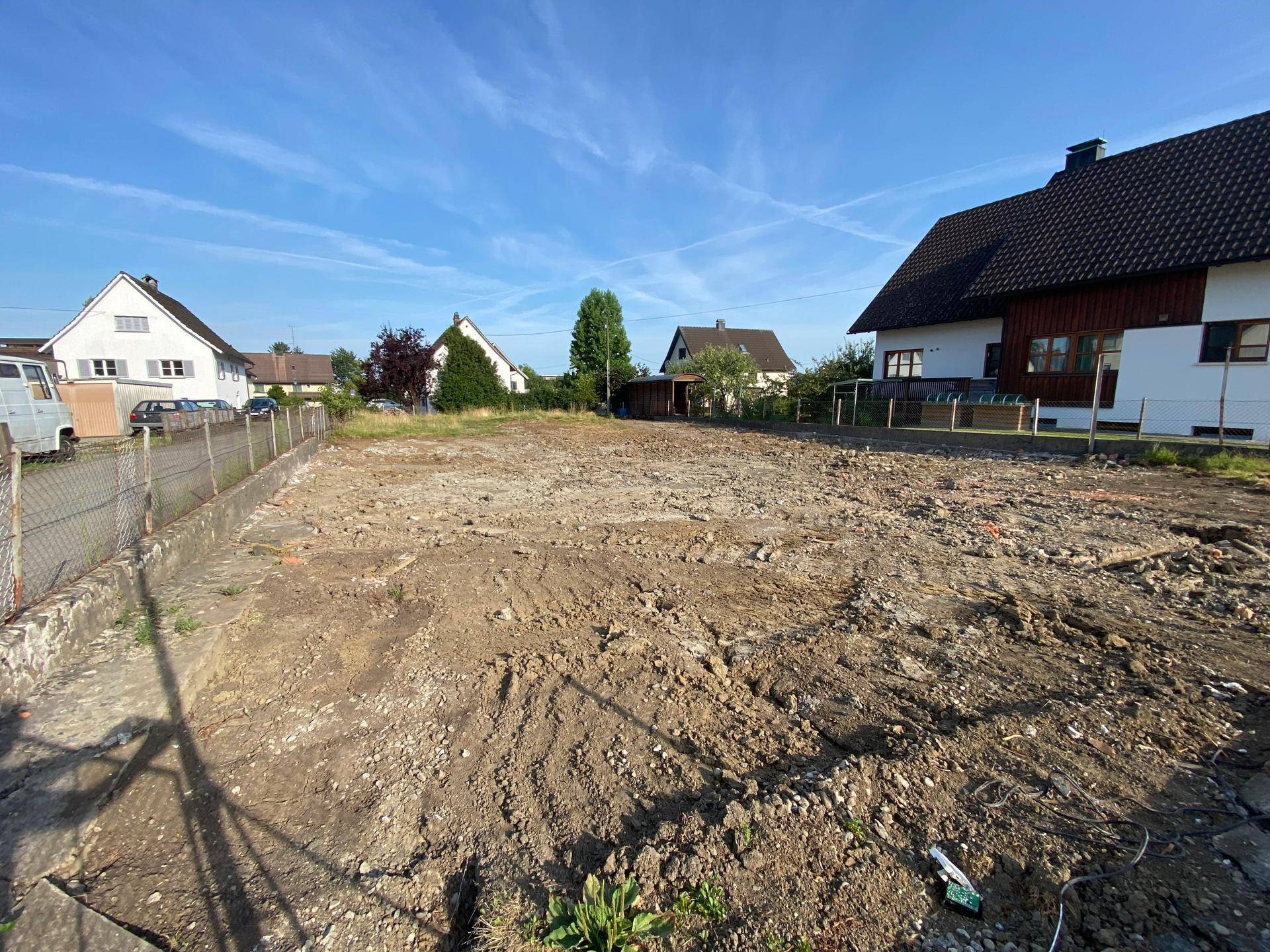 Freies Baugrundstück mit Erdboden und vereinzelten Pflanzen, umgeben von Wohnhäusern unter klarem Himmel.