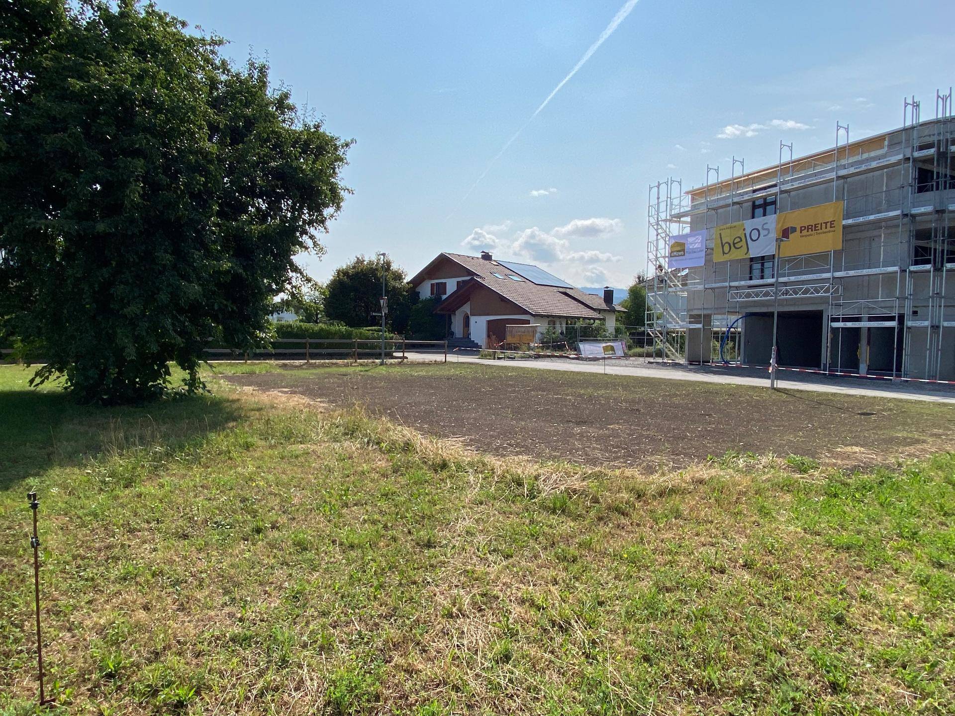 Unbebautes Grundstück mit Baustelle im Vordergrund und einem Wohnhaus mit Bergblick im Hintergrund.