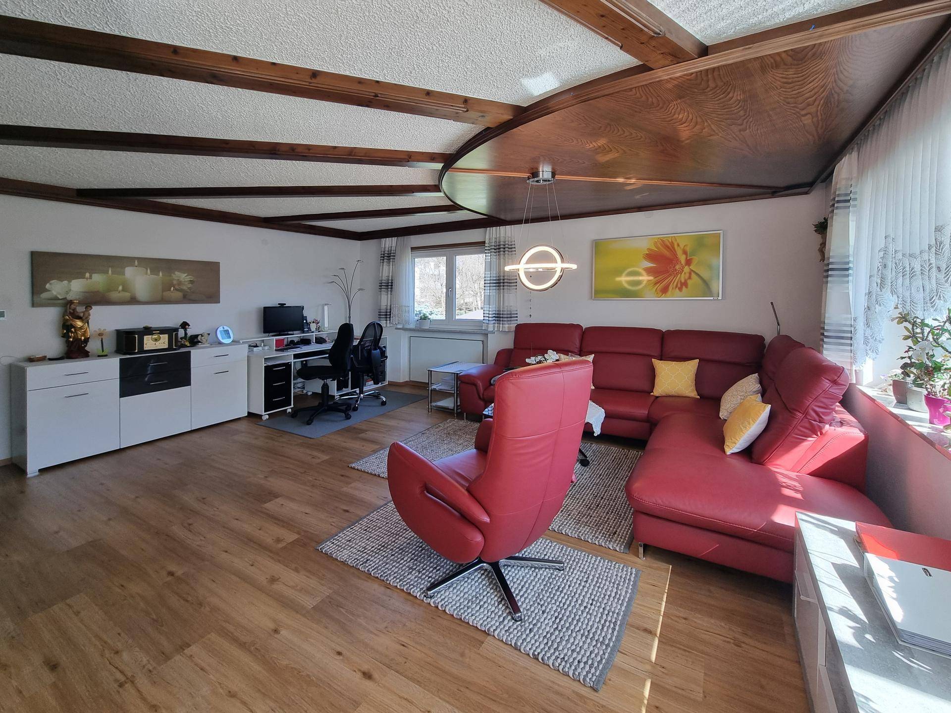 Gemütliches Wohnzimmer mit Holzbalkendecke, roter Ledercouch und Arbeitsbereich.